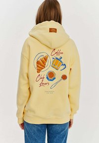 Žltá mikina s grafikou na chrbte: kanvica na kávu, croissant, šálka a lyžica s textom. Ťažká tkanina, uvoľnený strih, kapucňa s šnúrkou.