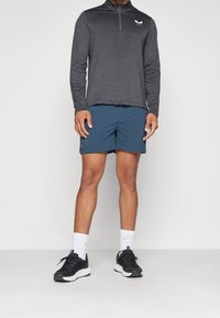 Urheiluasu, joka koostuu tummanharmaasta pitkähihanaisesta paidasta, jossa on vetoketjullinen kaulus ja logo. Yhdistetty sinisiin shortseihin ja mustiin, valkoisin yksityiskohdin varustettuihin juoksukenkiin.