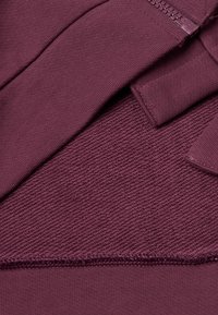 Pull en bordeaux avec fermeture éclair. Il présente un tricot lisse et un tissu texturé. Comprend un ourlet avec couture visible et des poignets lisses.