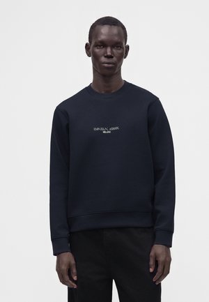Homme à la peau foncée portant un sweat-shirt noir Emporio Armani Milano et un pantalon noir, debout devant un fond clair uni.