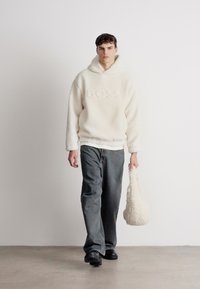 Bijela flis dukserica s utisnutim "BOSS" logom, u kombinaciji sa sivim farmerkama i pahuljastom bijelom torbicom na ramenu. Model nosi crne cipele.