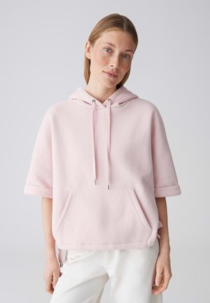 Frau mit hellbraunen Haaren trägt einen hellrosa kurzärmligen Hoodie und weiße Hosen, steht vor einem schlichten hellgrauen Hintergrund.