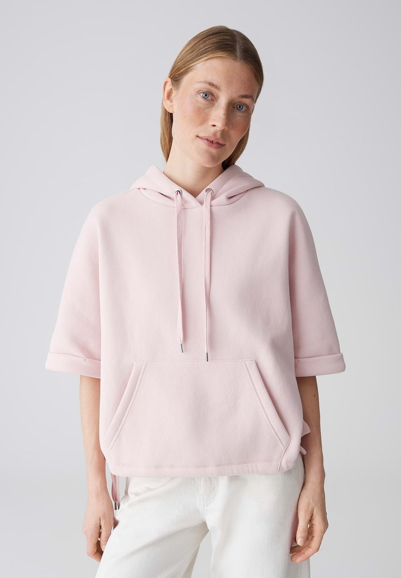 Frau mit hellbraunen Haaren trägt einen hellrosa kurzärmligen Hoodie und weiße Hosen, steht vor einem schlichten hellgrauen Hintergrund.