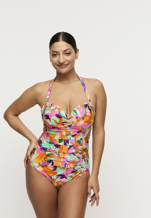 Vrouw met donker haar in een kleurrijk bloemen halterbadpak, staand met één hand op haar heup, glimlachend tegen een effen studiobackground.