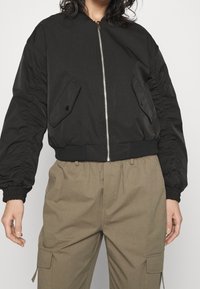 Černá bomber bunda se zipem vp�ředu, elastickými manžetami a dvěma předními kapsami. Vyrobena z lehkého materiálu s hladkou texturou.