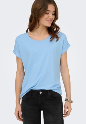 Joven mujer con una camiseta azul clara con las mangas remangadas, ligeramente metida dentro de unos vaqueros negros, con una pulsera de plata en su muñeca derecha.