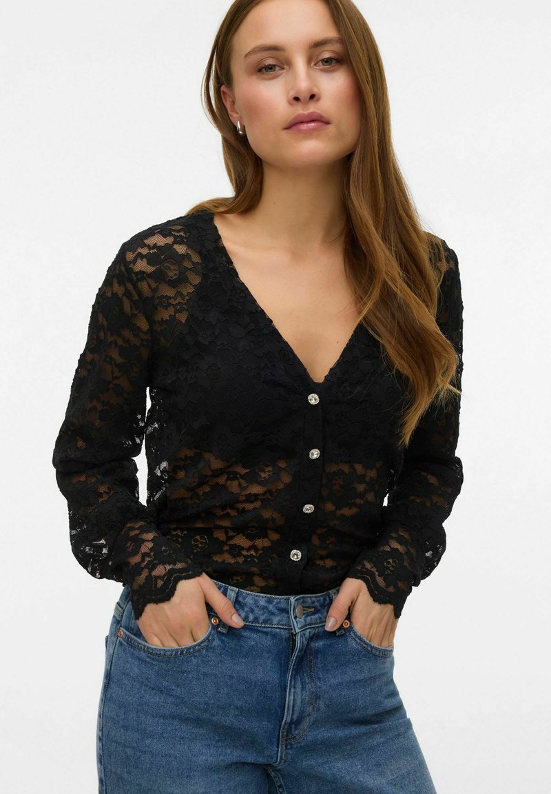 Cardigan nero in pizzo a maniche lunghe con scollo a V, decorato con bottoni e polsini smerlati, indossato sopra jeans blu a vita alta.