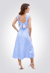 Vestido midi floral en azul claro con tirantes volantes y detalle de lazo en la parte trasera. Tela fluida con cintura ajustada y abertura en la espalda. Sandalias de tacón.