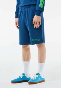 Pantalones cortos deportivos azules con una cinturilla elástica, que presentan un logo de Lacoste en verde y un emblema de cocodrilo. Combinados con zapatillas azules y calcetines blancos.