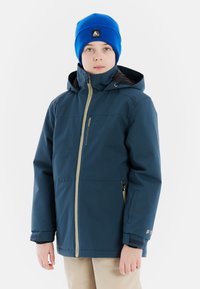 Marineblauwe waterdichte jas met capuchon, voorzien van een beige rits aan de voorkant en verstelbare manchetten, gecombineerd met een blauwe gebreide beanie.
