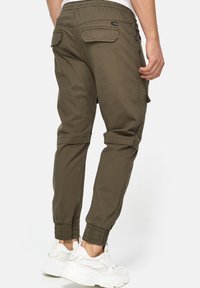 Olive green cargo spodenki z elastycznymi mankietami i wieloma kieszeniami, cechujące się teksturowanym materiałem i luźnym krojem. Stylizowane z białymi sneakersami.