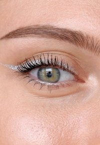 Close-up van een oog met een groene en bruine iris, lange zwarte wimpers en een zilveren eyeliner detail langs het ooglid. Glad huidoppervlak.