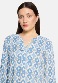 Blouse légère à motifs bleu et crème, présentant des formes géométriques et un col en V avec un détail accentué doré. Manches longues, coupe décontractée.