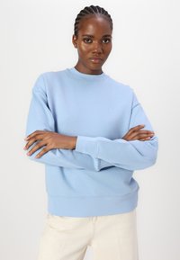 Felpa azzurra chiara con scollo rotondo, polsini e orlo a coste. Presenta un piccolo logo sul petto. Indossata con pantaloni di colore chiaro.