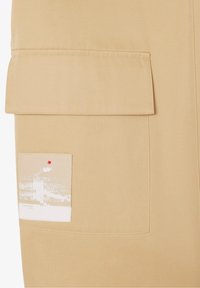 Tissu beige avec une grande poche à rabat. Une poche plus petite présente une impression blanche subtile et un point d'accent rouge. Texture lisse, design structuré.