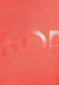 Korala tkanina ma gładką fakturę z dużymi, błyszczącymi literami tworzącymi napis "KO". Tło jest jednolite, co zwiększa widoczność tekstu.