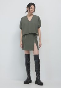 Top corto verde oliva con maniche a sbuffo, abbinato a una gonna mini coordinata con zip frontali e spacchi alti; stivali neri fino al ginocchio.