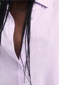 Chemise en lin lavande avec un col décontracté, présentant une texture subtile et des boutons transparents, partiellement déboutonnée, dévoilant un arrière-plan texturé.