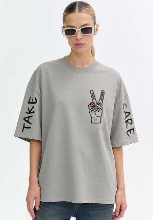 Vrouw draagt lichtgrijze oversized T-shirt met vrede-hand grafiek en woorden "FAITH LOVE PEACE JOY HOPE," "TAKE" op linkermouw, "CARE" op rechtermouw.
