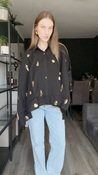 Camisa oversized preta com padrões de folha dourada, frente abertura com botões e mangas longas, combinada com jeans flare azuis claros.