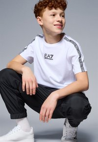 Camiseta blanca de manga corta con el logotipo de EA7, combinada con pantalones negros ajustados y zapatillas blancas. Tejido suave, diseño minimalista y puños elásticos.