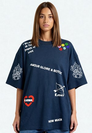 Femme aux cheveux bruns et raides portant un t-shirt oversize bleu marine avec des symboles et textes blancs et colorés, notamment "Karma," "Lover," et "Amour Gloire & Botox."
