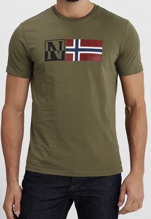 Mężczyzna ubrany w oliwkowozieloną koszulkę z czarnym napisem "N" i grafiką norweskiej flagi na piersi, w zestawieniu z ciemnymi dżinsami.