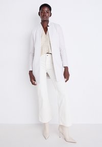 Cappotto lungo grigio chiaro su un top in maglia beige, abbinato a pantaloni a gamba larga crema e décolleté nudi con punta affusolata. Colori solidi, design elegante.