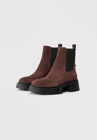 Botine din suede maro cu panouri elastice negre pe laterale și tălpi groase din cauciuc negru, așezate pe un fundal gri deschis.