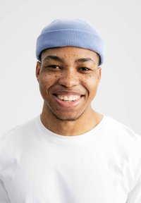 Lichtblauwe beanie, gemaakt van katoen, met ribbelstructuur. Het model draagt een wit t-shirt, glimlachend, met een zachte grijze achtergrond.