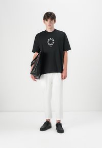 Czarna bawełniana koszulka z białym okrągłym logo, luźny krój, w połączeniu z białymi cropped spodniami i czarnymi butami na slip-on. Trzyma czarną skórzaną torbę.