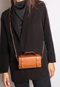 Borsa a mano in finta pelle marrone con manico superiore e hardware dorato, forma rettangolare e tracolla a catena, indossata sopra un blazer nero.