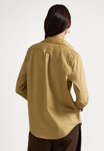 Žena s dlhými tmavými vlasmi, oblečená v béžovej košeli na gombíky a tmavých nohaviciach, stojaca chrbtom k jednoduchému bielemu pozadiu.