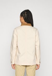 Persoon met gevlochten haar die een beige lange mouw jacket met een bruine kraag en beige broek draagt, staande met het gezicht naar een effen lichte achtergrond.