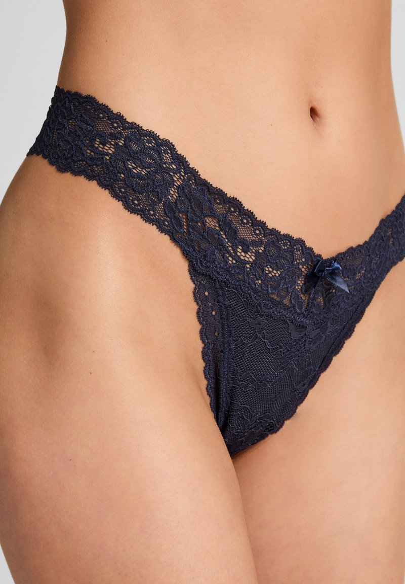 Marine kanten string met complexe bloemenpatronen, gegolfde randen en een kleine satijnen strikaccent aan de voorkant. Zachte stofstructuur.