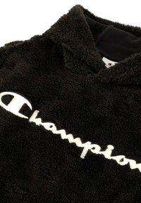 Sudadera de forro polar negra con capucha, que presenta un gran logo bordado en blanco de "Champion" en la parte frontal. Tejido suave y texturizado.