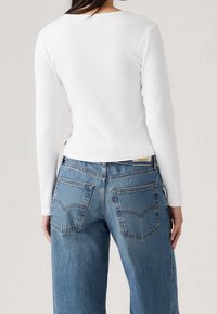 Bílý žebrovaný top s dlouhým rukávem a kulatým výstřihem, spojený s volnými modrými džínami s kapsami na zadní straně a prošitými detaily.