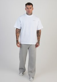 Mężczyzna z tatuażami, ubrany w białą koszulkę SikSilk, szare dresy i białe trampki, stojący na tle bez wzorów.