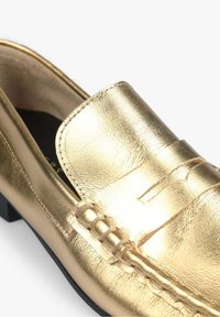 Mocassin en cuir doré avec une finition texturée, présentant un devant cousu et un dessus lisse, reposant sur une semelle noire et fine.