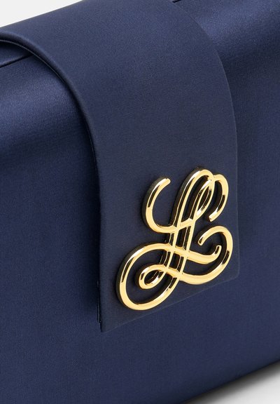 Marineblå håndtaske med en gylden dekorativ lås, der har et elegant, sammenflettet kursivdesign på frontklappen.