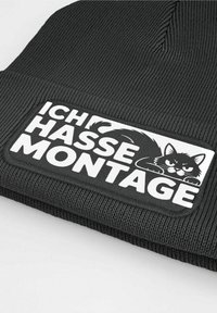 Black knit beanie with a ribbed texture and a rectangular patch featuring a graphic of a black cat and the text "ICH HASSE MONTAGE" in white.