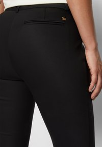 Gros plan d'une personne portant un pantalon noir ajusté avec un petit logo doré près de la poche arrière et une main visible reposant sur le côté.