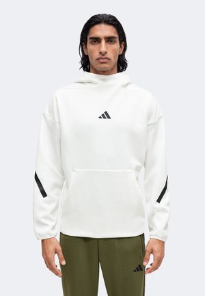 Mand med mørkt hår iført hvid Adidas-hættetrøje med sorte striber på ærmerne og olivengrønne Adidas-bukser mod en ensfarvet baggrund.