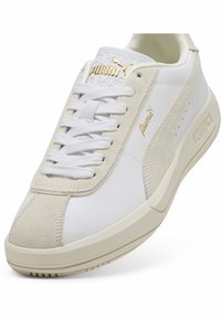 Baskets blanches avec détails en daim beige, dotées d'une semelle en caoutchouc, d'un système de laçage et d'un logo Puma doré sur le côté et la languette.