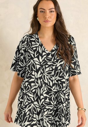 Femme aux longs cheveux bruns portant des boucles d'oreilles dorées, un haut à col en V noir et blanc à motif de feuilles, et un bracelet doré, debout devant un fond uni.