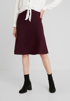 Vrouw met een donkerpaarse rok tot op de knieën, een witte blouse met strik en zwarte enkellaarsjes met hak, staand met één hand op haar heup.