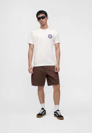 Joven con gafas de sol negras, camiseta blanca con gráfico, pantalones cortos marrones, calcetines blancos y zapatillas negras, de pie sobre un fondo liso.