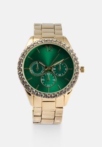 Montre-bracelet dorée avec un cadran vert, trois sous-cadrans et une lunette ornée de strass. Bracelet en métal avec une texture brossée.