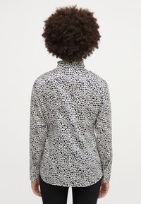 Chemise à manches longues en tissu de couleur claire avec un motif à pois noirs, dotée d'un col montant et d'une silhouette ajustée.