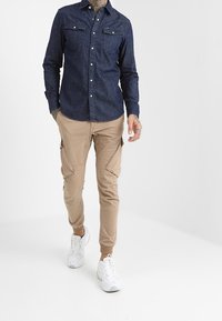 Mörk denimskjorta med två bröstfickor och silverfärgade knappar, matchad med beigefärgade cargobyxor med sidfickor och vita sportskor.